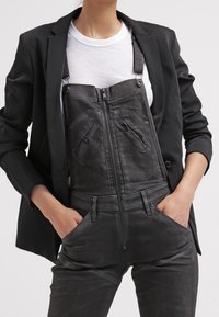 Black denim overalls with a zippered front, adjustable straps, and multiple zippered pockets, layered over a white T-shirt and black blazer.