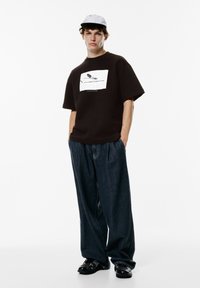 Mørkebrun oversized t-shirt med grafisk print på forsiden, parret med løstsiddende blå jeans og sorte sko. Lys grå cap.