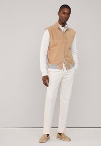 Chaleco de ante color marrón con cremallera, cuello de punto gris, suéter blanco de canalé debajo, pantalones blancos ajustados y zapatos beige tipo slip-on.