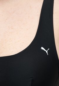Tissu de sport noir sur l'épaule avec logo Puma blanc visible sur une peau lisse avec de légères taches de rousseur.
