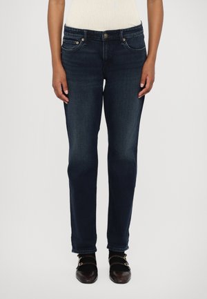 Mørkeblå denimjeans med rett passform, med frontlommer, knappestolpe og subtile sømdetaljer. Brukt med brune sko.