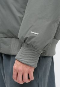 Graue Steppjacke mit gerippten Bündchen, mit dem weißen "DRYVENT"-Logo. Glatte Textur, teilweise zugeknöpft, an einem entspannten Ärmel präsentiert.