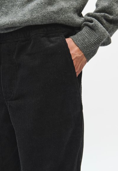 Personne portant un pantalon en velours côtelé noir avec la main dans la poche et un pull gris à manches longues avec des poignets côtelés visibles.