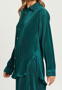 Camicia teal a coste realizzata in un tessuto lucido, con colletto, bottoni frontali e maniche a vestibilità ampia con polsi texturizzati. Orlo asimmetrico.