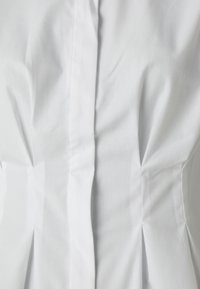 Bluză albă din bumbac cu un design ajustat, pliuri verticale și un guler clasic cu nasturi. Textură netedă, fără motive vizibile.