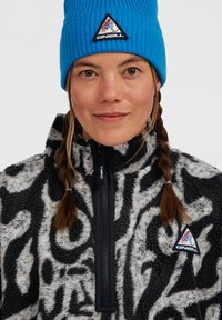 Jonge vrouw met gevlochten haar die een blauwe O'Neill-beanie en een zwart-wit gebreide fleecjas met O'Neill-logo's draagt.