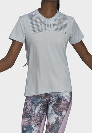Kvinde iført lysegrå kortærmet skjorte med perforeret øvre frontpanel og mønstrede lilla og blå leggings, armene let hævet.