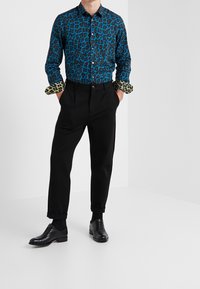 Blauwe en zwarte panterprint shirt met contrasterende panterprint manchetten, gecombineerd met zwarte geplooide broeken en zwarte leren schoenen.