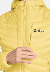 Mano che regola la zip della tasca sul petto di una giacca trapuntata gialla Jack Wolfskin con cappuccio.