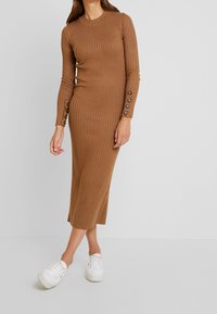 Abito a maglia marrone a coste con maniche lunghe, dettagli con bottoni ai polsini, lunghezza midi, indossato con sneakers bianche. Texture liscia e design aderente.