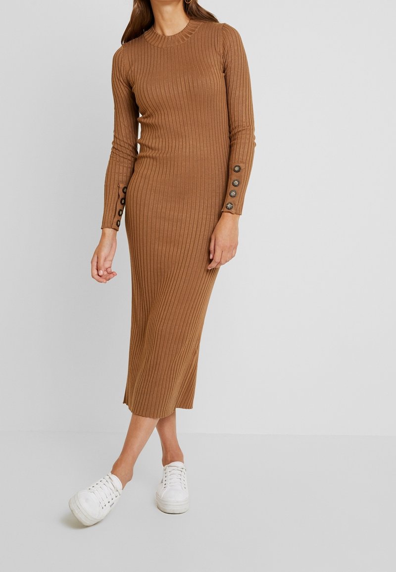 Abito a maglia marrone a coste con maniche lunghe, dettagli con bottoni ai polsini, lunghezza midi, indossato con sneakers bianche. Texture liscia e design aderente.