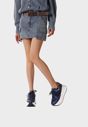 Donna che indossa gonna e camicia di denim blu, cintura marrone e sneakers platform blu navy, in piedi con la gamba destra piegata.