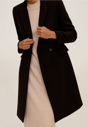Personne portant un manteau noir croisé sur une robe beige côtelée à col roulé, avec plusieurs bagues aux deux mains.