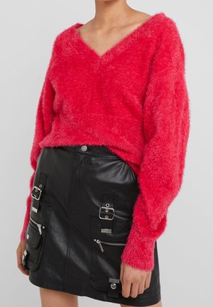 Femme portant un pull rouge vif en V en tissu duveteux et une mini jupe en cuir noir avec plusieurs boucles et poches zippées.