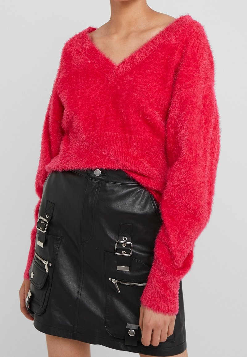 Femme portant un pull rouge vif en V en tissu duveteux et une mini jupe en cuir noir avec plusieurs boucles et poches zippées.