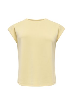 Femme souriante, portant un débardeur jaune pâle, un pantalon noir large et des chaussures pointues foncées, debout, les mains derrière le dos.