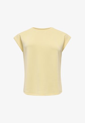 Femme souriante, portant un débardeur jaune pâle, un pantalon noir large et des chaussures pointues foncées, debout, les mains derrière le dos.