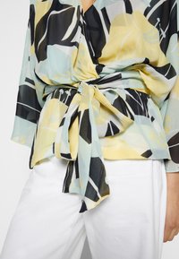 Femme portant une blouse à motifs avec des formes abstraites jaunes, noires et bleu pastel nouée à la taille, associée à un pantalon blanc.