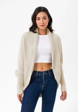 Beige gestrickte Strickjacke mit gerippter Textur, breitem Schalkragen und überschnittenen Schultern, kombiniert mit einem weißen, kurz geschnittenen Oberteil und dunklen Jeans.