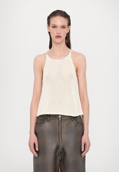 White, knitted halter top with a loose fit, textured pattern, and thin straps, paired with dark brown, high-waisted pants with zipper details.