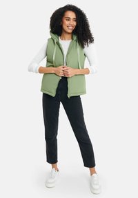 Gepolsterte grüne Weste mit Kapuze, vorderem Reißverschluss und Kordelzügen, getragen über einem weißen Langarmshirt und kombiniert mit schwarzen Hosen und weißen Sneakern.