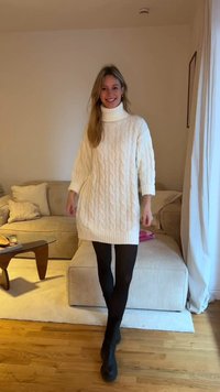 White cable-knit oversized sweater dress with a high collar; paired with black tights and ankle boots. Textured, soft finish.