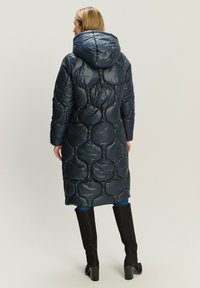 Marineblaue Steppjacke mit einem gepolsterten, bubbelförmigen Design. Verfügt über eine große Kapuze, lange Ärmel und reicht bis zur Mitte der Wade. Kombiniert mit Jeans und schwarzen kniehohen Stiefeln.