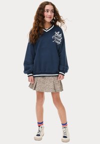 Sweat-shirt bleu marine avec le texte blanc "Anti Social Mood", col rayé ; associé à une jupe évasée à imprimé léopard. Baskets blanches avec des rayures colorées.