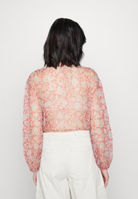 Donna con capelli scuri di media lunghezza, che indossa un top lungo a maniche trasparenti con stampa floreale e pantaloni bianchi a vita alta, in piedi di spalle.