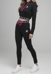 Hoodie noir et joggeurs avec des cordons et des marquages rose. Tissu lisse, design ajusté, et baskets blanches complètent la tenue.