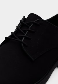 Μαύρο δετό παπούτσι με απαλή υφή, στρογγυλεμένη μύτη και αντίθετες ραφές κατά μήκος των ακμών. Απλός, κομψός σχεδιασμός.