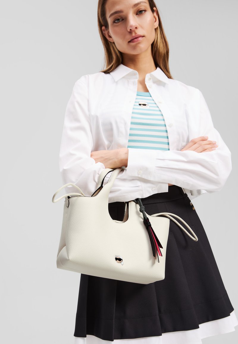 White leather handbag with two handles, featuring a textured finish and decorative tassels in red and black. Flat, open zipper design.