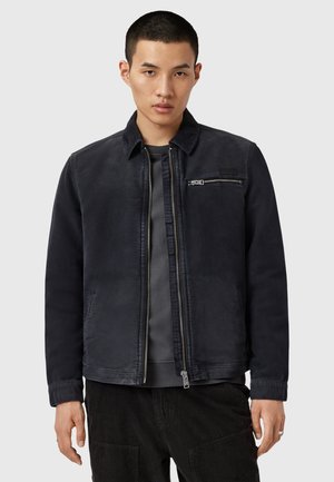 Giacca in cotone nero misto con zip frontale, colletto a punta, tasca sul petto e tasche laterali. Tessuto liscio con dettagli di design minimi.