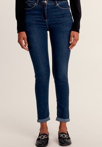 Slim-fit, donkerblauwe denim jeans met een hoge taille, opgerolde pijpen en contrasterende goudkleurige stiksels. Draagt het samen met zwarte instapschoenen met hardware.