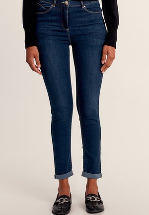 Slim-fit, donkerblauwe denim jeans met een hoge taille, opgerolde pijpen en contrasterende goudkleurige stiksels. Draagt het samen met zwarte instapschoenen met hardware.