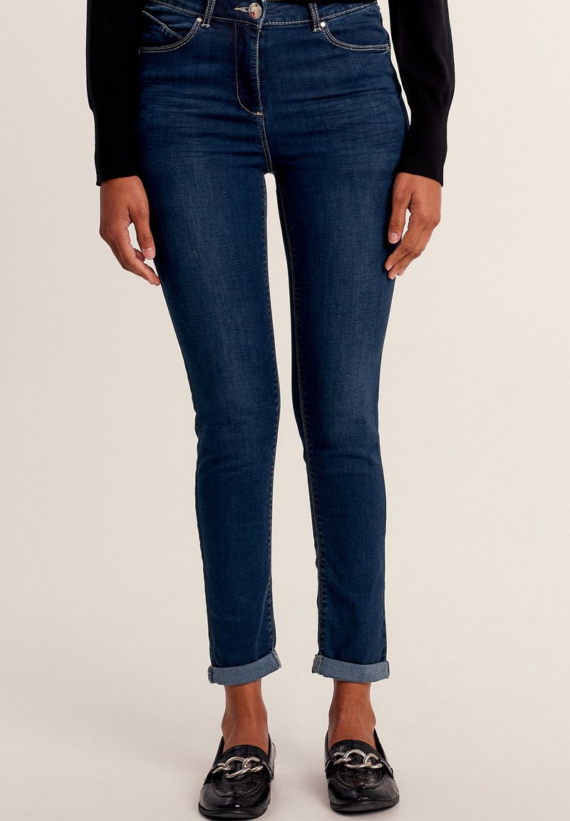 Slim-fit, donkerblauwe denim jeans met een hoge taille, opgerolde pijpen en contrasterende goudkleurige stiksels. Draagt het samen met zwarte instapschoenen met hardware.