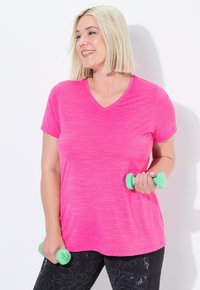 Donna con capelli biondi a caschetto, che indossa una maglietta rosa e leggings neri con un motivo, mentre tiene in mano due manubri verde chiaro, sorridendo e guardando a destra.