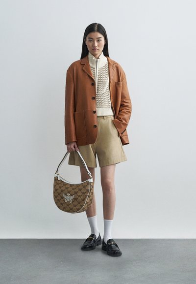 Egy nő barna bőrkabátot, krémszínű kötött pulcsit, bézs rövidnadrágot, fehér zoknit és fekete loafert visel, miközben egy mintás barna kézitáskát tart.