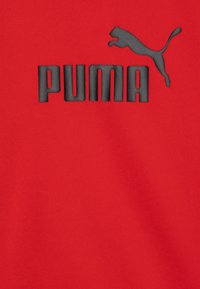 Sarkans audums izceļas ar izteiktu melnu "PUMA" logotipu, virs kura atrodas lēkājoša puma siluets, demonstrējot gludu tekstūru un drosmīgu dizainu.
