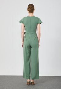 Grön overall med korta ärmar, figurnära midja, vida byxor och dragkedja bak. S jämn textur, minimalistisk design och inga mönster.