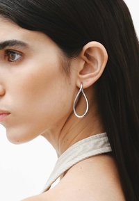 Pendientes en forma de lágrima plateados cuelgan de la oreja, con un acabado suave y un diseño minimalista y elegante. Se puede ver una cinta de tela texturizada.
