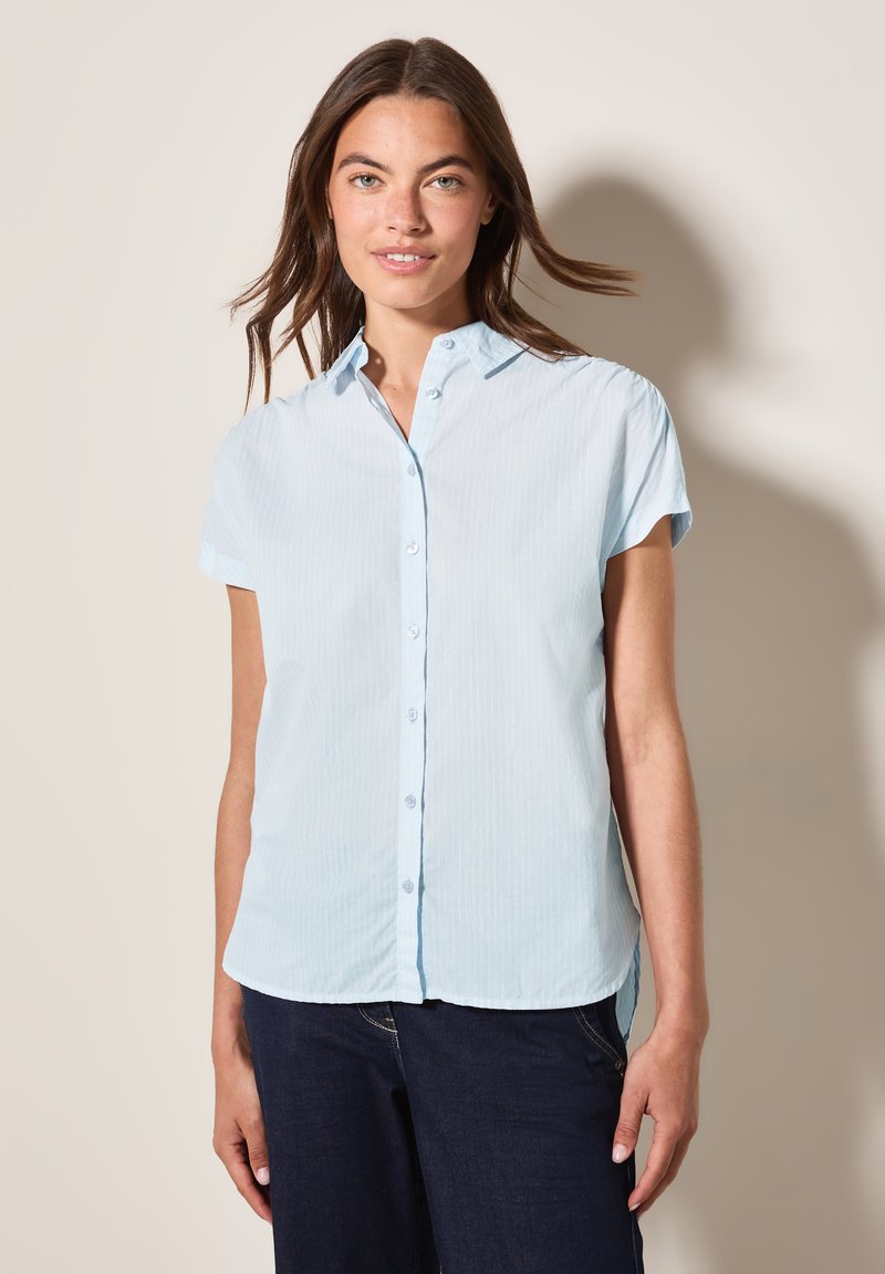 Chemise à manches courtes bleu clair, avec un col et un devant à boutons, dotée d'une texture rayée douce. L'ourlet se situe au niveau des hanches.