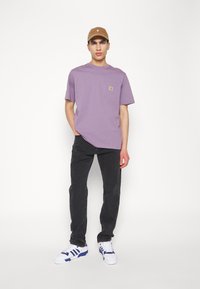 Moški v rjavi kapici, sivem kratkoročnem srajcu s sprednjim žepom, črnih kavbojkah in belih športnih copatih z modrimi črtami stoji ob belem ozadju.