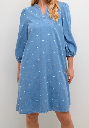 Femme portant une robe bleue mi-longue aux genoux avec des manches 3/4 bouffantes et de petits motifs de cœurs blancs, debout devant un fond uni.