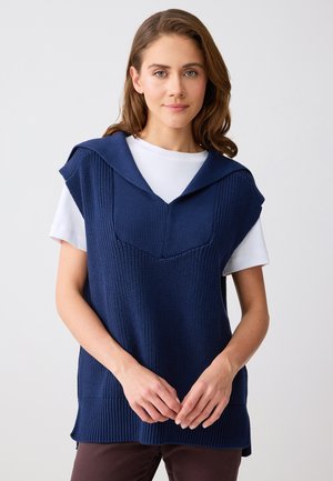 Vrouw met lichtbruin haar, gekleed in een marineblauw gebreid vest over een witte korte mouwen shirt, staand tegen een effen lichte achtergrond.