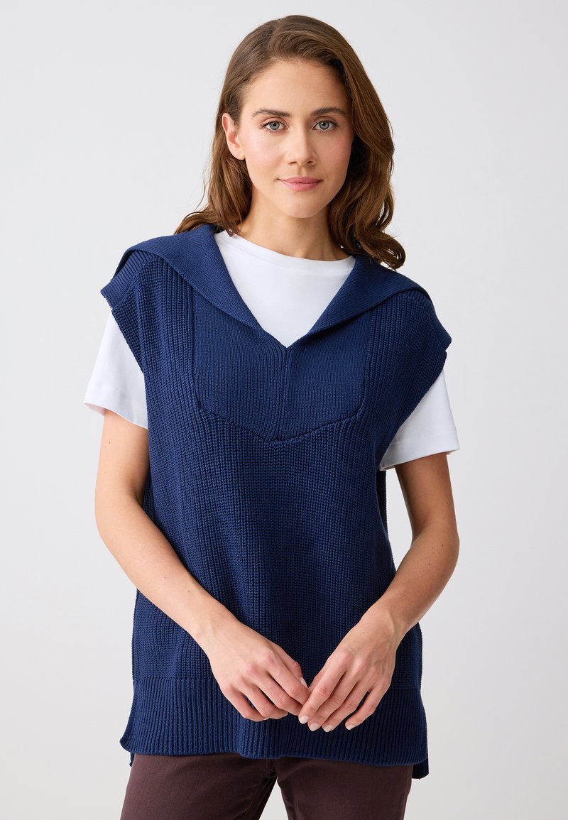 Femme aux cheveux châtain clair portant un gilet en maille bleu marine par-dessus une chemise blanche à manches courtes, debout devant un fond clair uni.