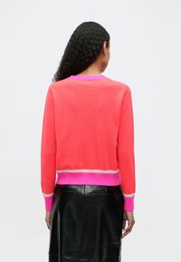 Mujer con cabello rizado largo viste un suéter coral con borde beige y rosa brillante, combinado con una falda de cuero negra, de espaldas sobre un fondo blanco.