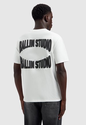 Jonge man van achteren gezien, die een wit T-shirt draagt met grote zwarte tekst "BALLIN STUDIO" die twee keer op de rug gedrukt is.