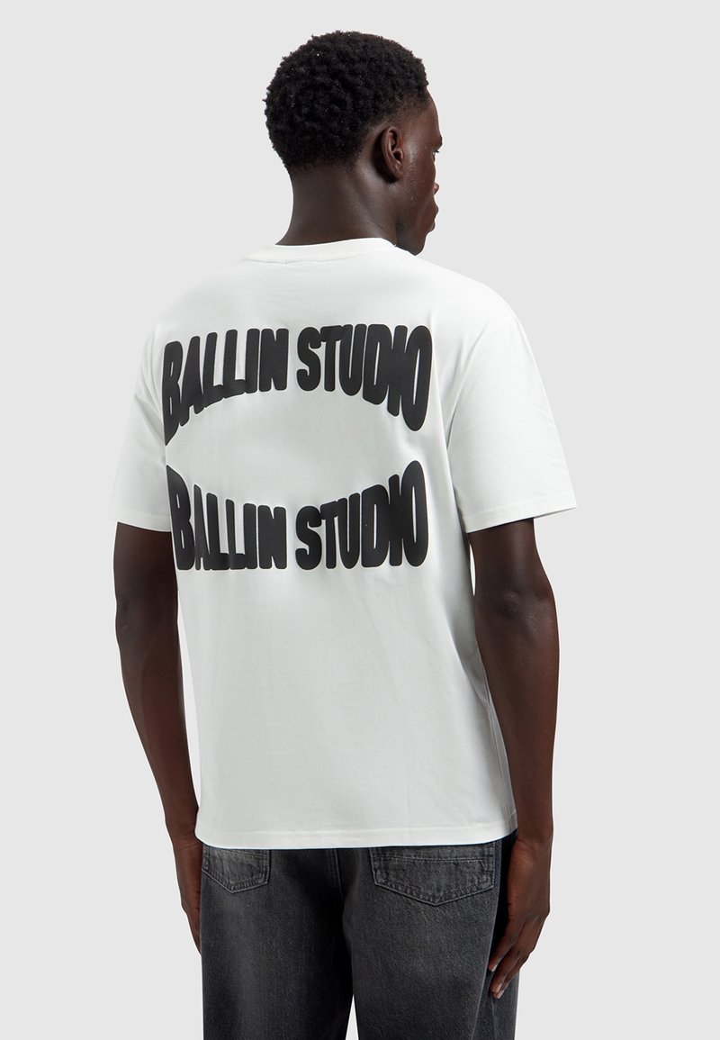 Jonge man van achteren gezien, die een wit T-shirt draagt met grote zwarte tekst "BALLIN STUDIO" die twee keer op de rug gedrukt is.