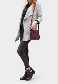 Szary sweterek o fakturze, czarna koszula w wzory, ciemne dopasowane dżinsy i czarne botki za kostkę z paskami; nosi bordową torbę na ramię.